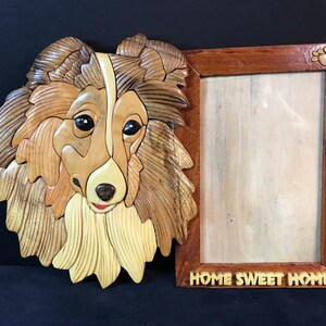 May include: A wooden picture frame with a carved wooden dog head on the left side. The frame has a light wood finish and the dog head is a light brown color. The frame has the words "Home Sweet Home" carved into the bottom.
