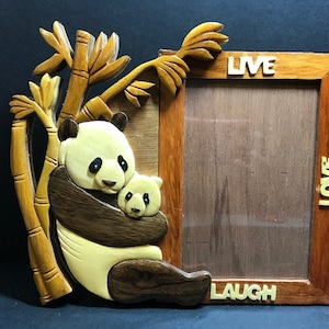 Może przedstawiać: Drewniana ramka na zdjęcia z rzeźbionym pandą i wzorem bambusa. Ramka zawiera słowa "LIVE", "LOVE" i "LAUGH". Panda obejmuje małą pandę. Ramka jest wykonana z drewna.