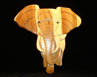 May include: A wooden carving of an elephant, with a light brown color and dark brown wood grain. The elephant's trunk is long and curved, and its eyes are inlaid with black.