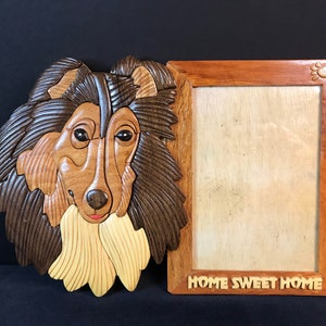 May include: A wooden picture frame with a carved collie dog head on the left side. The frame has a light wood finish and the words "Home Sweet Home" are carved into the bottom.