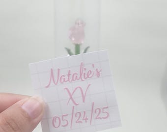 May include: A clear champagne flute with the text "Natalie's XV 05/24/25" in pink script. A small pink rose is visible inside the glass. A square sticker with the same text is held in front of the glass.