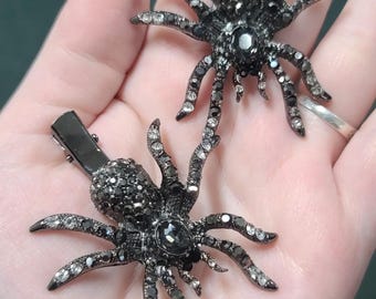 May include: Two black spider hair clips, each with a detailed design. The spiders have multiple legs and are embellished with small, sparkling crystals. The clips are held in a hand against a dark green background.