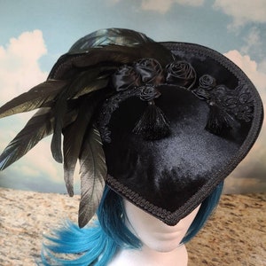 May include: A black velvet heart-shaped hat with black feathers and black satin roses. The hat has a black trim and is worn on a mannequin with blue hair.