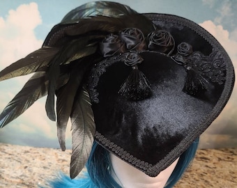 May include: A black velvet heart-shaped hat with black feathers and black satin roses. The hat has a black trim and is worn on a mannequin with blue hair.