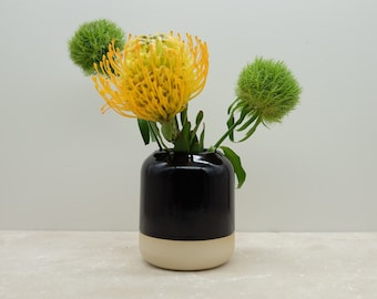 Könnte beinhalten: Eine schwarze und weiße Keramikvase mit abgerundeter Form, die gelbe und grüne Blumen hält.