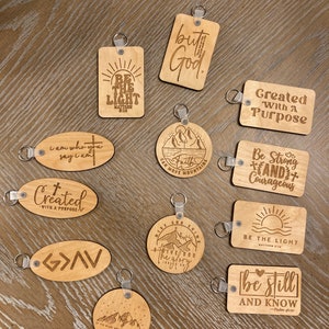 May include: A collection of 11 wooden keychains with inspirational messages and mountain scenes. The keychains are engraved with phrases like "Be the Light", "Created with a Purpose", "Be Still and Know", and "Rise and Shine Give God the Glory".