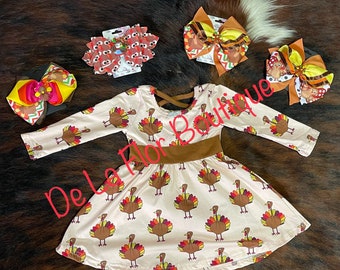 May include: A light beige dress with a brown waistband and long sleeves, patterned with cartoon turkeys. Several hair bows with turkey and fall-themed designs are also displayed. The text "De La Flor Boutique" is visible.
