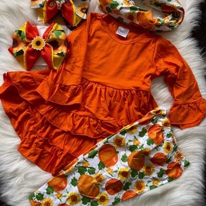 May include: A long-sleeve orange tunic with a ruffled hem, white leggings featuring a pumpkin and sunflower print, a matching white headband with pumpkins and sunflowers, and two hair bows with orange, yellow, and sunflower patterns.