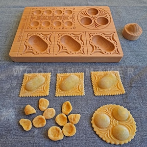 Puede incluir: Un molde para pasta rectangular de madera clara con hendiduras talladas para dar forma a la masa, con el texto WOODEN MAKER ITALIA grabado en el lateral. Junto a él hay varias piezas de masa de pasta cruda moldeadas, incluyendo tres raviolis cuadrados, una pieza redonda con múltiples huecos y varias formas pequeñas tipo orecchiette.
