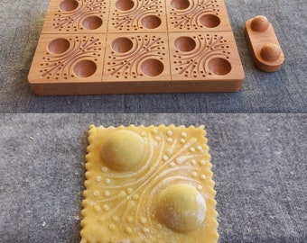 May include: A wooden ravioli mold with nine individual sections, each with a decorative design. The mold is used to create patterned ravioli. A separate wooden piece with two round indents is also included. The mold is shown with a finished ravioli, featuring a decorative design and two filled pockets.