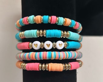 May include: A set of five colorful beaded bracelets with gold accents. The bracelets are stacked on a black display stand. Three of the bracelets have white beads with gold heart charms.