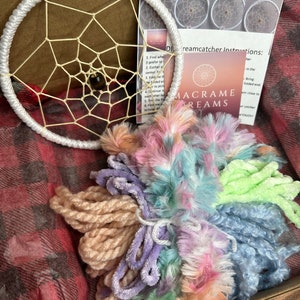 May include: A DIY dreamcatcher kit in a box. The kit includes a white-wrapped circular frame with a woven web design, colorful yarn, and instructions. The yarn colors include pastel pink, blue, green, and purple. The kit is labeled "MACRAME DREAMS".