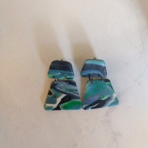 Puede incluir: Un par de pendientes hechos a mano con un diseño veteado. Los pendientes tienen una forma rectangular en la parte superior y trapezoidal en la inferior. Los colores incluyen tonos de azul, verde, negro y blanco. Los pendientes están conectados con elementos metálicos plateados.