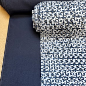 May include: A roll of navy blue fabric with a white geometric pattern. The fabric is a dark blue color and has a smooth texture. The pattern is a repeating design of connected circles.