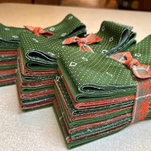 May include: Stacks of green and orange cloth napkins with a white diamond pattern. Each stack is tied with a decorative orange ribbon. The napkins are folded and stacked neatly, creating a visually appealing arrangement.