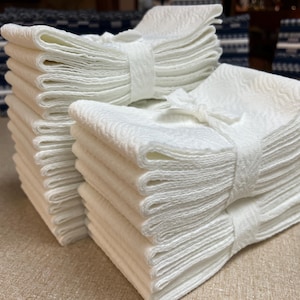 May include: Several stacks of white, textured cloth napkins tied with matching fabric bands. The napkins are neatly folded and arranged, creating a clean and organized presentation. The background features other folded textiles.