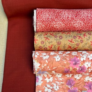 May include: A selection of fabric rolls in various patterns and colors. The fabrics include a solid deep red, a red and white geometric pattern, a tan floral print, and two floral prints in shades of peach and pink.