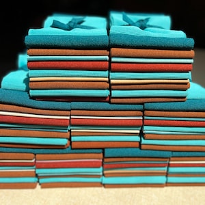 May include: Stacks of folded fabric in various colors, including teal, rust, and brown. Some stacks are topped with teal bows. The fabrics appear to be of a textured material, possibly towels or linens, arranged in neat piles.