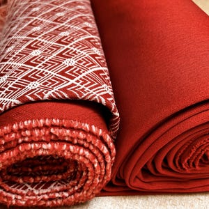 May include: Two rolls of fabric, one patterned with a white geometric design on a red background, and the other a solid red. The patterned fabric is partially unrolled, revealing its texture. The solid fabric is tightly rolled.