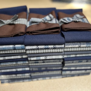 May include: A stack of folded fabric items in shades of blue and brown. The top layer features three sets of folded brown fabric with a bow detail, resting on a layer of solid navy blue fabric. Below are stacks of patterned and solid blue fabrics.