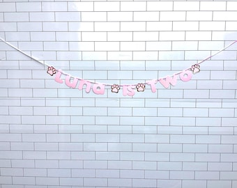 May include: A decorative banner featuring light pink cutout letters that spell out the phrase LUNA IS TWO, interspersed with small rose gold paw print accents, all strung together on a thin white ribbon for hanging display.