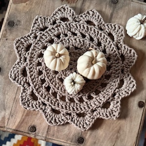 Könnte beinhalten: Beige gehäkelte Tischdecke mit gewelltem Rand, verziert mit vier kleinen, weißen Kürbissen. Die Tischdecke liegt auf einer rustikalen Holzoberfläche und erzeugt eine herbstliche Ästhetik. Die Kürbisse variieren in Größe und Form.