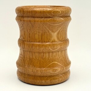 May include: A handcrafted wooden vase with a light brown finish. The vase features a detailed, carved design with curved lines and circular accents. The vase has a cylindrical shape with a wider base and top, and is approximately 5 inches tall.