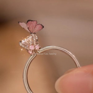 Może przedstawiać: Srebrny pierścionek z przezroczystym kamieniem szlachetnym i zawieszką w kształcie różowego motyla. Pierścionek jest ozdobiony małymi różowymi kamieniami szlachetnymi wokół kamienia.