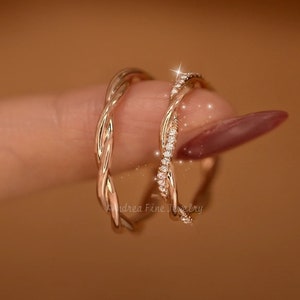 Könnte beinhalten: Zwei goldfarbene Ringe, einer mit einem gedrehten Design und der andere mit einem gedrehten Design und einer Reihe kleiner, klarer Steine. Die Ringe befinden sich an einem Finger.