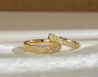 May include: Two gold wedding bands with a textured bark-like design. The rings are displayed on a white surface with a neutral background. The bands are likely made of gold and are designed for a wedding or engagement.