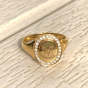 以下が含まれることがあります： 縁に小さな透明な石が並んだ、透明な楕円形の石がセットされた金のシグネットリング。石の中央に「M」の文字が刻まれています。