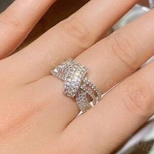 以下が含まれることがあります： 結び目のデザインと透明なキュービックジルコニアの石が付いた銀の指輪。指輪は人の指にはめられています。