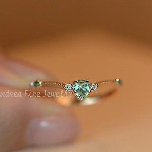 May include: A delicate gold ring with a small green gemstone and tiny diamonds set in a curved band. The ring is on a white surface.