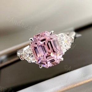 Könnte beinhalten: Ein Verlobungsring aus Weißgold mit einem rosafarbenen Smaragdschliff und zwei kleineren Diamanten an jeder Seite des Mittelstücks.