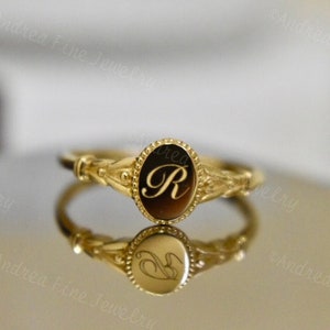 Puede incluir: Un anillo de sello de oro con un bisel de forma ovalada que presenta la letra "R" grabada en su interior. El anillo tiene un diseño delicado y adornado con un borde de cuentas.