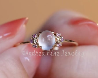 May include: A delicate gold ring with a round, milky white gemstone surrounded by smaller pink gemstones. The ring is on a finger with light pink nail polish.