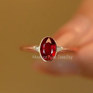 May include: A silver ring with a red oval gemstone and two smaller diamonds on either side.  ©Andrea Fine Jewelry