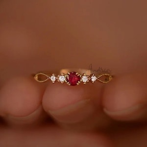 May include: A delicate gold ring with a red gemstone surrounded by smaller clear gemstones.