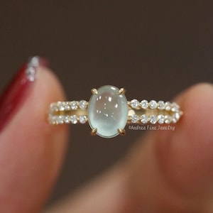 Puede incluir: Un anillo de oro con una piedra preciosa ovalada de color verde claro, flanqueada por dos filas de pequeñas piedras preciosas transparentes. El anillo tiene un diseño delicado, con las palabras "Andrea Fine Jewelry" visibles en la banda.