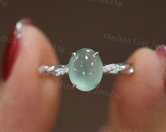 May include: A silver ring with a light green oval gemstone. The band has a twisted design with small, clear accent stones. The ring is held up by a person with red nail polish.