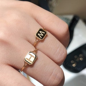 Puede incluir: Dos anillos de oro con esmalte negro y letras grabadas. El anillo de la izquierda tiene la letra "H" y el anillo de la derecha tiene la letra "M".