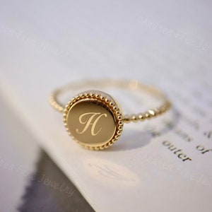 May include: A gold ring with a round, beaded bezel featuring the letter "H" engraved in the center. The ring is on a white surface.