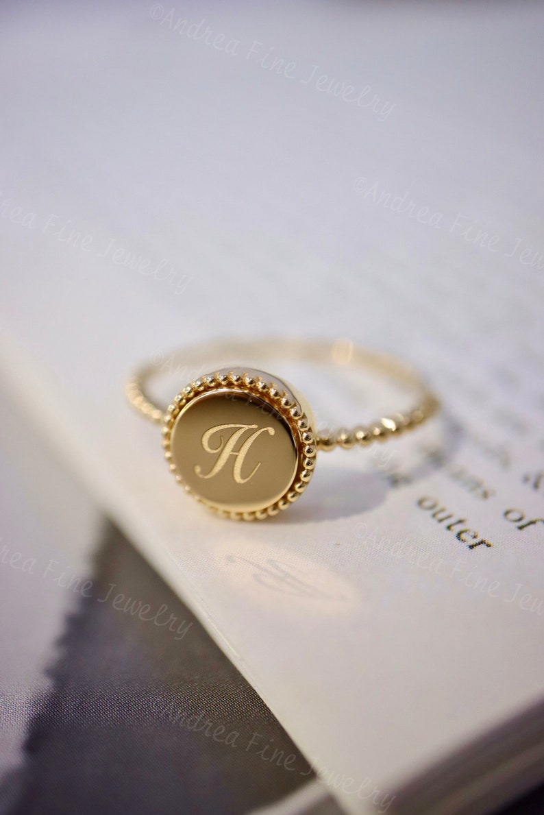 May include: A gold ring with a round, beaded bezel featuring the letter "H" engraved in the center. The ring is on a white surface.