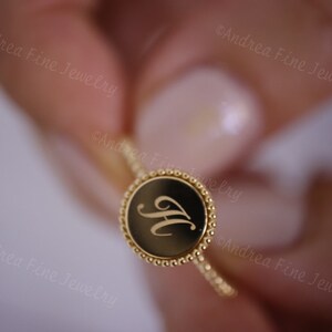 May include: A gold ring with a round, black, engraved disc featuring the letter "H" in a cursive font.