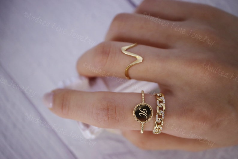 May include: A close-up of a hand wearing three gold rings. The ring on the index finger is a simple band with a small round charm that has the letter "H" engraved on it. The ring on the middle finger is a thin band with a delicate chain design. The ring on the ring finger is a simple, curved band.