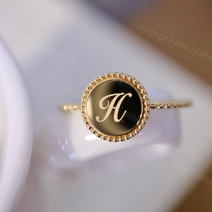 May include: A gold ring with a round bezel featuring the letter "H" engraved in a cursive font. The bezel is surrounded by a row of small beads.