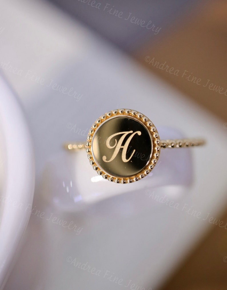 May include: A gold ring with a round bezel featuring the letter "H" engraved in a cursive font. The bezel is surrounded by a row of small beads.