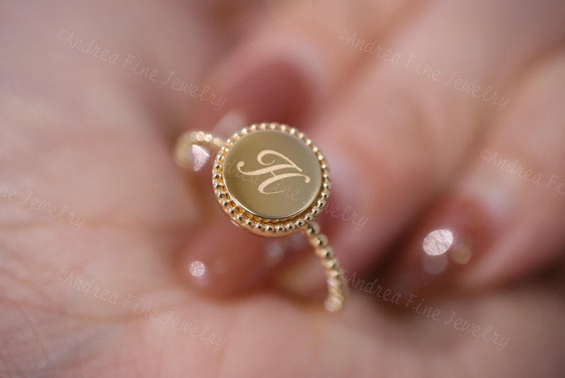 May include: A gold ring with a round, beaded bezel featuring the letter "H" engraved in a cursive script.