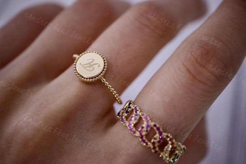 May include: Two gold rings. The first ring has a round, gold, engraved plate with the letter "M" on it. The second ring is a chain link style with multi-colored gemstones.