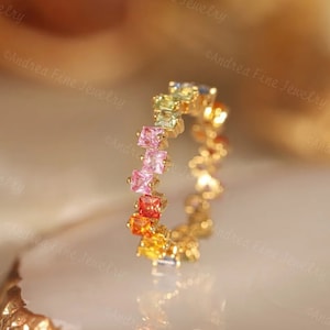 Op de afbeelding: Een gouden bandring met een regenboog van vierkante edelstenen. De ring is bezet met roze, oranje, gele, groene en blauwe stenen.